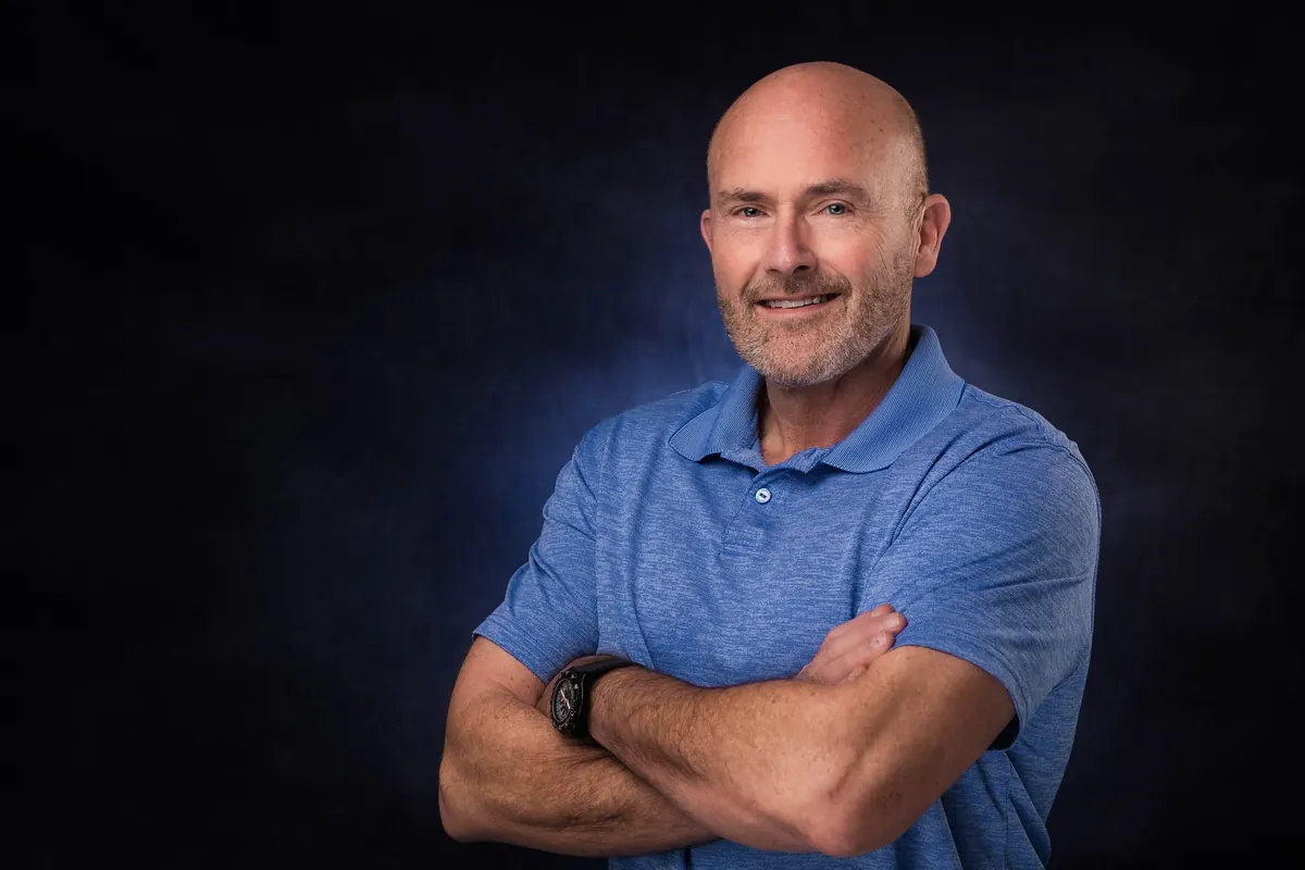 Studio headshot of a smiling man in a blue polo with arms crossed on a dark blue mottled backdrop