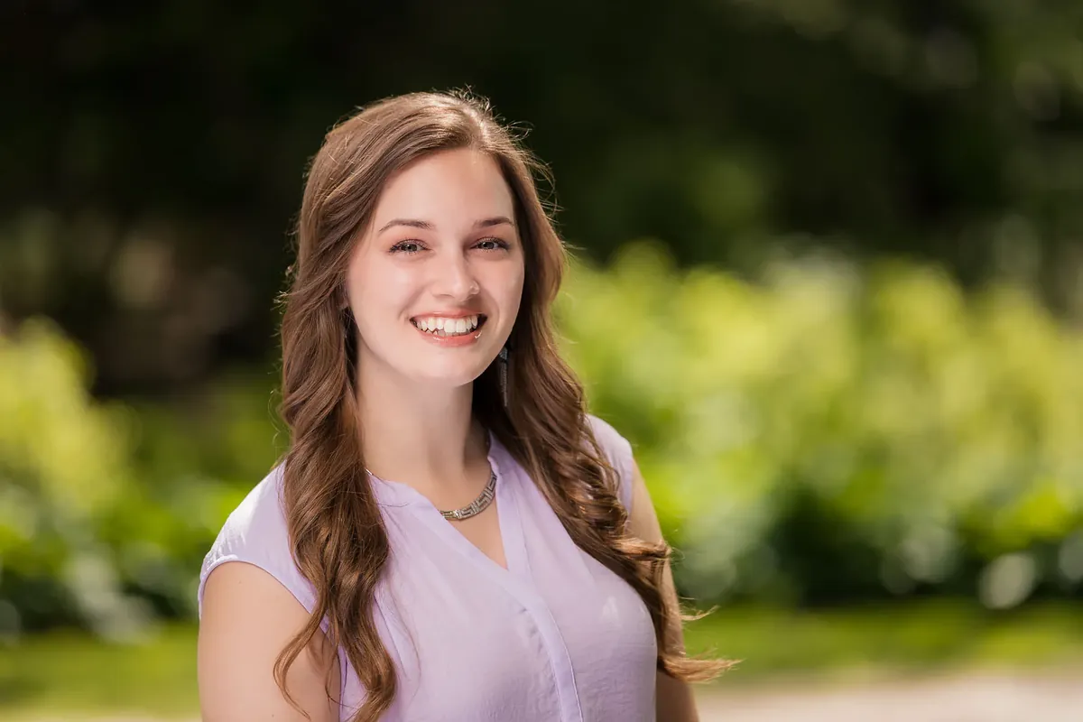 Outdoor headshot of a smiling young woman with long brown hair in a lavender top, natural light with soft green background