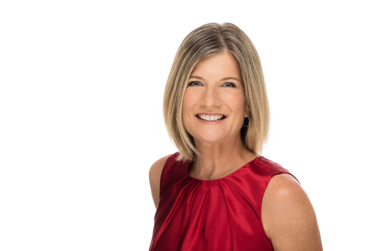 Professional headshot of a smiling woman with a blonde bob in a red satin top, photographed on a white seamless studio backdrop