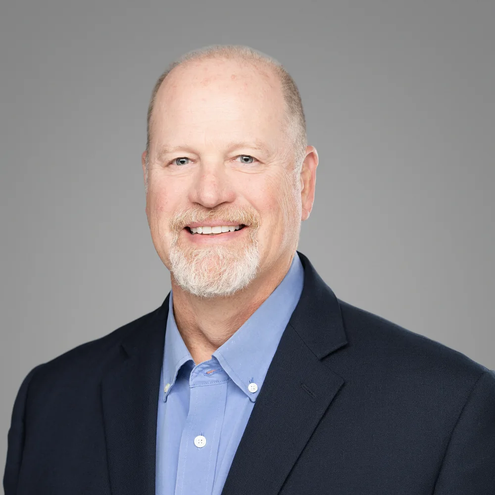Professional headshot of a smiling man with a white goatee in a navy blazer and light blue shirt on a gray backdrop