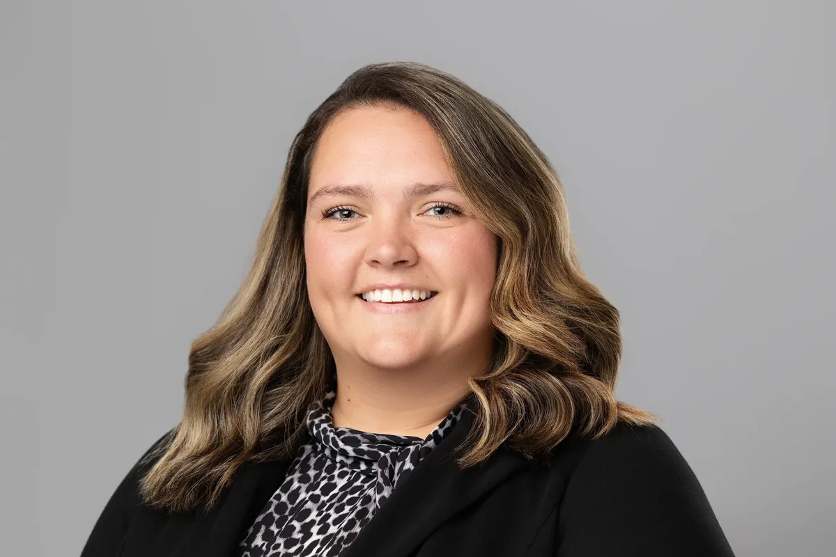 Professional headshot of a smiling woman with wavy highlighted hair in a black blazer and patterned blouse on a gray studio backdrop